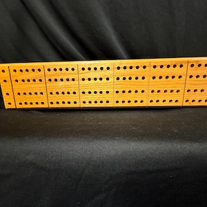 May include: A wooden cribbage board in a light brown hue. It features rows of holes for pegging, arranged in a rectangular layout with a subtly angled end. This board is designed for the card game of cribbage.