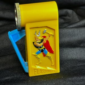 May include: A yellow Flashmite flashlight featuring a blue handle and a silver-colored bezel. The front of the flashlight has a raised image of Thor, a superhero, holding a hammer. The word "FLASHMITE" is printed below the image.