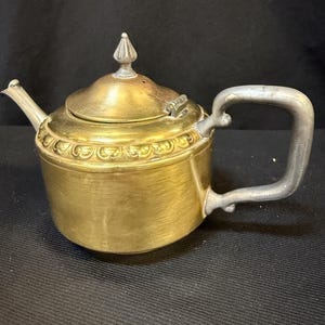 Könnte beinhalten: Eine Vintage-Teekanne aus Messing mit einem silbernen Griff und Ausguss. Die Teekanne hat einen abgerundeten Körper, einen Deckel mit dekorativer Spitze und ein verziertes Design um die Basis des Deckels. Der Griff hat eine gebogene, rechteckige Form.