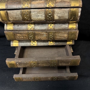Puede incluir: Una caja de madera dise&ntilde;ada para parecerse a una pila de libros antiguos. La caja es marr&oacute;n con detalles met&aacute;licos dorados en el lomos y las esquinas. Dos cajones est&aacute;n parcialmente abiertos, revelando el interior.