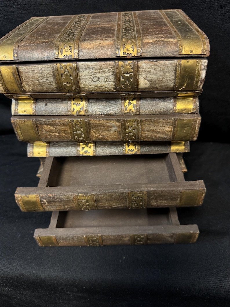 Puede incluir: Una caja de madera dise&ntilde;ada para parecerse a una pila de libros antiguos. La caja es marr&oacute;n con detalles met&aacute;licos dorados en el lomos y las esquinas. Dos cajones est&aacute;n parcialmente abiertos, revelando el interior.