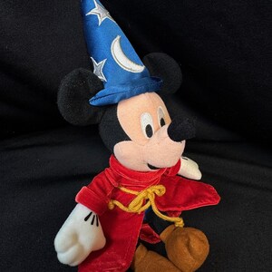 K&ouml;nnte beinhalten: Ein Pl&uuml;sch-Mickey Mouse als Zauberlehrling verkleidet. Die Figur tr&auml;gt einen blauen Zauberhut mit silbernen Sternen und einem Mond, einen roten Samtumhang und braune Schuhe. Die Figur ist etwa 25 cm gro&szlig;.