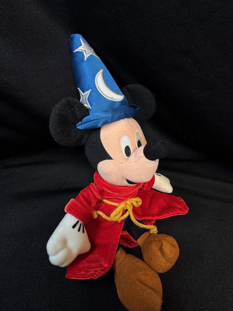 K&ouml;nnte beinhalten: Ein Pl&uuml;sch-Mickey Mouse als Zauberlehrling verkleidet. Die Figur tr&auml;gt einen blauen Zauberhut mit silbernen Sternen und einem Mond, einen roten Samtumhang und braune Schuhe. Die Figur ist etwa 25 cm gro&szlig;.