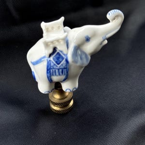 May include: A white and blue ceramic elephant figurine with a brass base. The elephant has blue accents on its ears, trunk, and saddle. The figurine is a decorative item, possibly a lamp finial or a small sculpture.