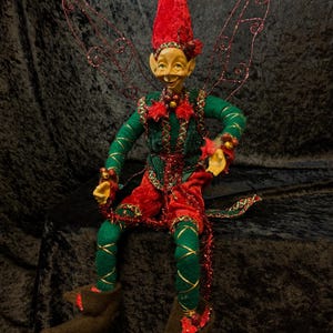Puede incluir: Una caprichosa figura de elfo con una cara sonriente, que lleva un gorro puntiagudo rojo y un atuendo verde y rojo. El elfo tiene delicadas alas de alambre y está adornado con detalles dorados. El elfo está sentado y tiene botas marrones.