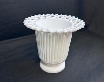 May include: A white, vintage-style vase with a fluted body and a decorative, openwork rim. The vase has a round base and stands on a dark background. The overall design is elegant and classic.
