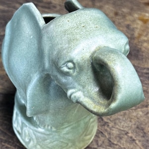 May include: A light green ceramic elephant-shaped pitcher. The pitcher features detailed facial features, including eyes and a trunk. The base has a decorative pattern. The pitcher is a unique decorative item.