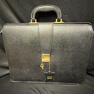 May include: Black leather briefcase with a top handle and gold-colored hardware. The briefcase has a flap closure with a key and a tag that reads "GENUINE LEATHER". The briefcase is rectangular and appears to be new.