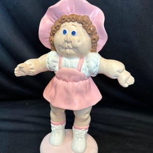 Puede incluir: Muñeca de cerámica vintage con sombrero rosa, vestido y zapatos. La muñeca tiene cabello rizado castaño, ojos azules y está de pie sobre una base circular rosa. Los brazos de la muñeca están extendidos.