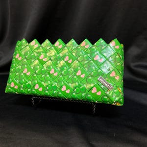 May include: A green wallet with a woven design, featuring pink hearts and the letter "m". The wallet has a rectangular shape and is displayed on a black surface. The brand name "ecotaf" is visible.