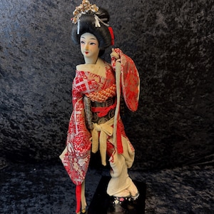 Peut inclure: Poupée japonaise de geisha avec une coiffure noire ornée de détails dorés et blancs. La poupée porte un kimono à motifs rouges et blancs avec de longues manches et une ceinture beige. La poupée est posée sur un socle noir.