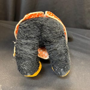 Puede incluir: Un juguete de peluche vintage con pelaje negro y detalles de lunares naranjas y blancos. Los pies del juguete est&aacute;n delineados en amarillo. El juguete est&aacute; sobre una superficie oscura.