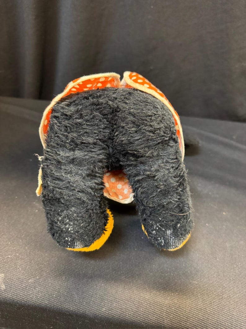 Puede incluir: Un juguete de peluche vintage con pelaje negro y detalles de lunares naranjas y blancos. Los pies del juguete est&aacute;n delineados en amarillo. El juguete est&aacute; sobre una superficie oscura.