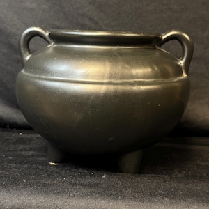 Puede incluir: Un caldero de cerámica negra con dos asas arqueadas y tres patas pequeñas. El caldero tiene un cuerpo redondeado y un acabado brillante, adecuado para uso decorativo o funcional.