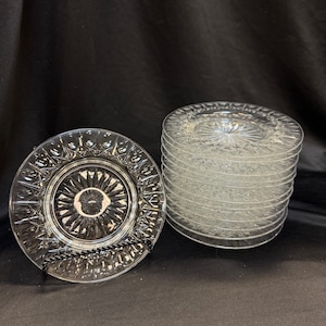 May include: Clear glass plates with a detailed, textured design. One plate is displayed on a black wire stand, while a stack of similar plates sits to the right. The plates have a circular pattern radiating from the center.
