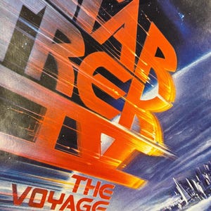May include: A movie poster close-up with the title "STAR TREK THE VOYAGE HOME" in orange and white lettering. The background features a space scene with a planet and a city.