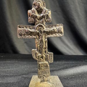 May include: A detailed silver-colored Orthodox cross with a figure of Jesus Christ. The cross is mounted on a square base. The design includes intricate carvings and religious iconography, set against a dark background.