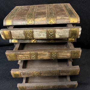 Puede incluir: Una caja de almacenamiento de madera con cuatro cajones extra&iacute;bles, dise&ntilde;ada para parecerse a libros antiguos apilados. La caja tiene un acabado marr&oacute;n desgastado con detalles decorativos de metal dorado en los bordes y lomos. Los cajones est&aacute;n abiertos.