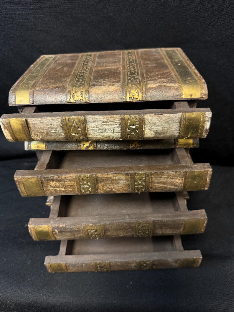 Puede incluir: Una caja de almacenamiento de madera con cuatro cajones extra&iacute;bles, dise&ntilde;ada para parecerse a libros antiguos apilados. La caja tiene un acabado marr&oacute;n desgastado con detalles decorativos de metal dorado en los bordes y lomos. Los cajones est&aacute;n abiertos.