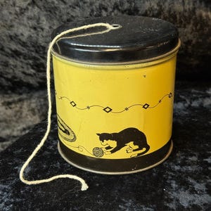 Puede incluir: Una lata amarilla vintage con tapa negra y cuerda adjunta. La lata presenta un gato negro jugando con una bola de hilo y un borde negro decorativo. La lata es cilíndrica y parece ser de metal.