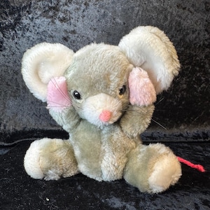Könnte beinhalten: Ein Stofftier, das eine graue Maus mit großen weißen Ohren und rosa Innenohren darstellt. Die Maus sitzt mit ihren Pfoten an den Ohren und hat einen roten Schwanz. Das Spielzeug befindet sich auf einem dunklen Hintergrund.