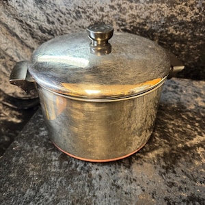 Pode incluir: Uma panela de cozinha em aço inoxidável com tampa e alça preta. A panela tem uma superfície brilhante e reflexiva e um botão preto na tampa. A parte inferior da panela tem um anel vermelho fino.