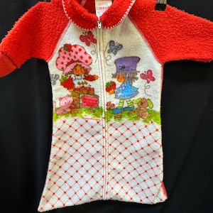 May include: A vintage red and white fleece jumpsuit featuring a Strawberry Shortcake graphic. The jumpsuit has a zip-up front, red sleeves, and a white body with a heart pattern. The collar is red with white trim.