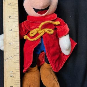 K&ouml;nnte beinhalten: Eine Pl&uuml;schpuppe von Mickey Mouse mit einem roten Samtmantel und einer goldfarbenen Krawatte. Die Puppe hat braune Schuhe, eine schwarze Hose und wei&szlig;e Handschuhe. Die Puppe ist etwa 28 cm gro&szlig;.