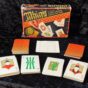 May include: Vintage "Mhing" card game box with cards. The box is red, black, and gold with the game name and illustrations. Several card stacks and individual cards are visible, featuring various symbols and characters.