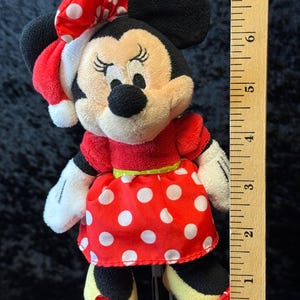May include: A Minnie Mouse plush doll, featuring a red dress with white polka dots, a matching bow, and yellow shoes. The doll has black ears and a red and white polka dot bow. The doll measures approximately 6 inches in height.