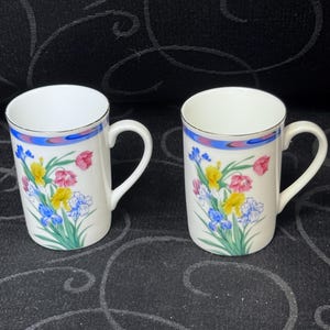 May include: Two white ceramic mugs with floral designs. Each mug features a gold rim, a blue and pink band near the top, and a colorful floral arrangement of pink, yellow, and blue flowers. The mugs have curved handles.