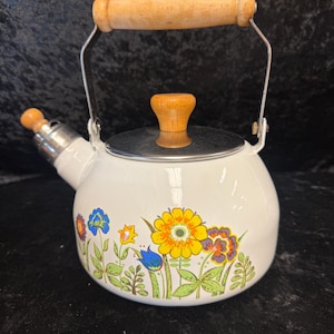 May include: White enamel tea kettle with a wooden handle and lid knob. The kettle features a floral design with yellow, blue, and orange flowers. The spout and handle supports are silver-colored.