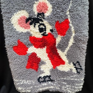 May include: A gray, rectangular, textured rug featuring a cartoon mouse design. The mouse has a red scarf, red heart-shaped hands, and a pink and white head. The rug is made of a soft, plush material.