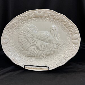 Puede incluir: Plato ovalado blanco roto con un diseño de pavo en relieve en el centro. El borde presenta motivos florales y de hojas. El plato mide aproximadamente 40 cm de largo.