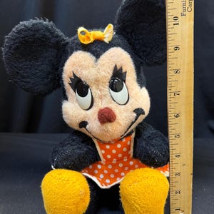 Puede incluir: Mu&ntilde;eca de peluche vintage de Minnie Mouse con orejas negras, cara melocot&oacute;n y nariz roja. Viste un vestido naranja con lunares blancos y tiene pies amarillos. Un lazo amarillo adorna su cabeza. La mu&ntilde;eca mide aproximadamente 25 cm de altura.