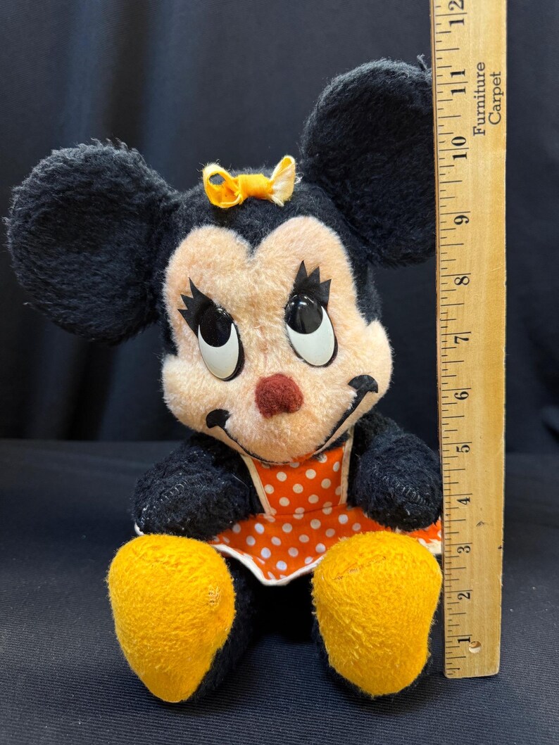 Puede incluir: Mu&ntilde;eca de peluche vintage de Minnie Mouse con orejas negras, cara melocot&oacute;n y nariz roja. Viste un vestido naranja con lunares blancos y tiene pies amarillos. Un lazo amarillo adorna su cabeza. La mu&ntilde;eca mide aproximadamente 25 cm de altura.