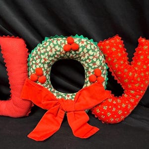 May include: A festive Christmas decoration spelling out "JOY". The letters are red, with a textured "J" and a "V" patterned with small flowers. A wreath with a red bow and holly accents completes the holiday display.
