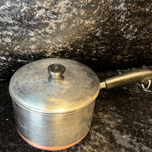 May include: A stainless steel saucepan with a lid and a black handle. The pot has a copper-coloured base and a black knob on the lid. The saucepan is designed for cooking and is suitable for various kitchen tasks.