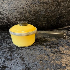 Puede incluir: Una pequeña cacerola amarilla con tapa y un solo asa. La olla tiene un borde plateado y un asa negra. La tapa es de color verde oscuro con un pequeño pomo redondo. La cacerola es probablemente vintage y adecuada para cocinar porciones pequeñas.