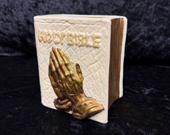 May include: A ceramic book-shaped container with the words "HOLY BIBLE" embossed on the front. The front of the container features a gold-coloured sculpture of praying hands. The book has a white exterior and brown-coloured edges.