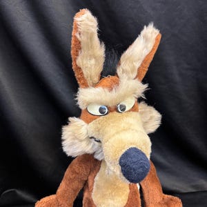 Può includere: Un peluche di Wile E. Coyote, con una combinazione di colori marrone e beige, un naso nero e grandi occhi espressivi. Il coyote ha lunghe orecchie soffici e un aspetto morbido e strutturato, tipico di un peluche.