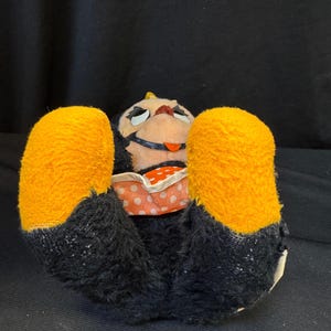 Puede incluir: Un juguete de peluche vintage en posici&oacute;n supina, con cuerpo negro, grandes pies naranjas y una cara pintada. El juguete lleva una prenda con estampado de lunares.