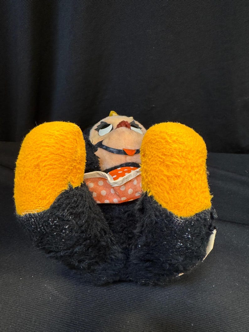 Puede incluir: Un juguete de peluche vintage en posici&oacute;n supina, con cuerpo negro, grandes pies naranjas y una cara pintada. El juguete lleva una prenda con estampado de lunares.