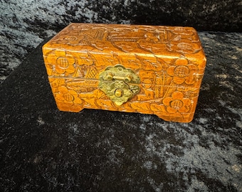 May include: A rectangular wooden box with intricate floral carvings and a brass clasp. The box is a warm, golden-brown color and sits on small feet. The carvings depict flowers and other decorative elements.