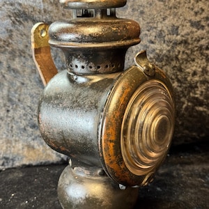 Puede incluir: Linterna antigua negra y bronce con una lente circular transparente. La linterna tiene un cuerpo redondeado, una parte superior con ranuras de ventilación y un asa. El metal muestra signos de edad y desgaste, con zonas de óxido y decoloración.