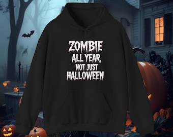 Pode incluir: Moletom preto com a frase branca "ZOMBIE ALL YEAR, NOT JUST HALLOWEEN". O moletom tem um bolso frontal e um capuz com cordão. O fundo apresenta uma cena assustadora de Halloween com morcegos, abóboras e uma casa mal-assombrada.