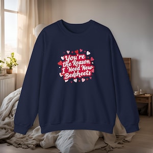 Könnte beinhalten: Marineblaues Sweatshirt mit dem Text "You're the Reason I Need New Bedsheets" in einem herzförmigen Design. Der Text ist rot und weiß, umgeben von roten und weißen Herzen. Das Sweatshirt ist ein lässiges Kleidungsstück.