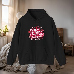 Könnte beinhalten: Schwarzer Kapuzenpullover mit Fronttasche und Kordelzugkapuze. Auf der Vorderseite befindet sich eine Grafik mit dem Text "You're the Reason I Need New Bedsheets", umgeben von roten und rosa Herzen. Der Hoodie wird auf einem Bett präsentiert.