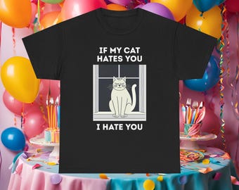 May include: Black t-shirt with the text "IF MY CAT HATES YOU I HATE YOU." The graphic shows a white cat in a window. The background includes colourful balloons and a decorated table, suggesting a party or celebration.
