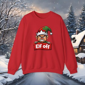 Peut inclure: Un sweat-shirt rouge avec un dessin d'elfe en colère portant un chapeau vert et rouge et tenant une pancarte « Elf off ». L'arrière-plan montre un paysage hivernal enneigé avec des arbres et une maison.